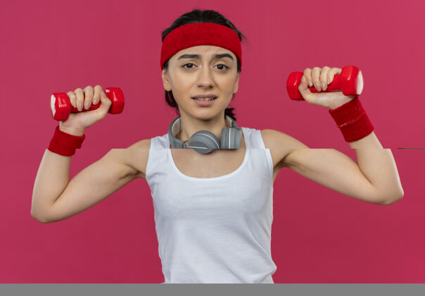 抱着身穿运动服的年轻健身女 头上带着两个哑铃 站在粉红色的墙上做着表情混乱的练习人女人哑铃