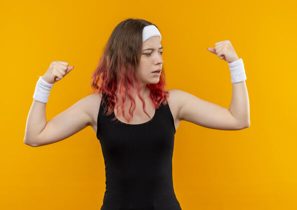 姿势身着运动服的年轻健身女士举起拳头 站在橙色的墙上 展现出自信的二头肌运动举起拳头