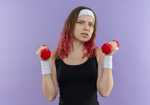 运动穿着运动服的年轻健身女士站在紫色的墙上用哑铃做练习 脸上带着恼怒的表情运动员烦恼女人