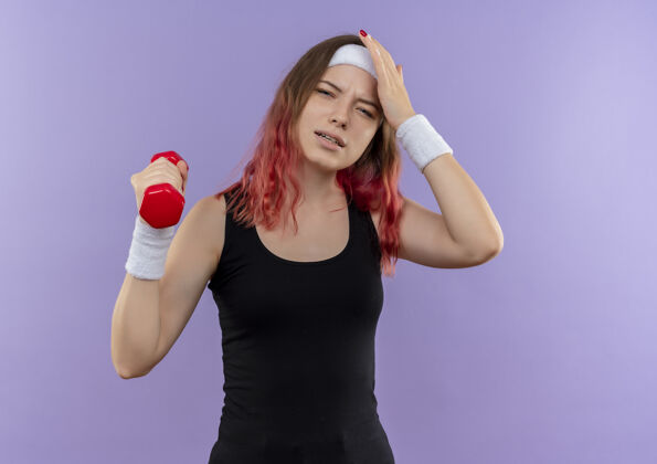 人身穿运动服的年轻健身女士举着哑铃站在紫色的墙上 看起来很累人运动累