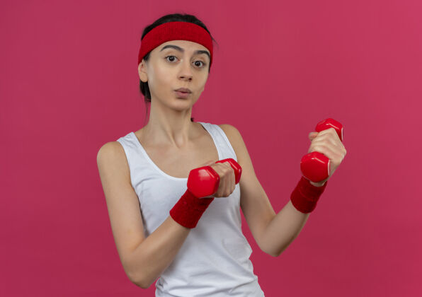 举起身穿运动服 头箍 双手举着两个哑铃 自信满满的年轻健身女士站在粉色的墙上做运动锻炼运动站着