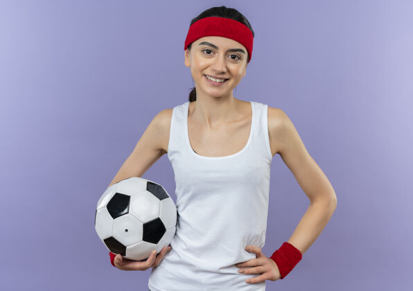 微笑身穿运动服 头箍 手持足球 微笑 自信 快乐 积极向上的年轻健身女士站在紫色的墙上年轻运动装自信