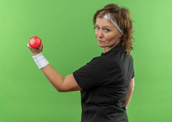 人中年运动女性 身穿黑色t恤 戴着头带 举着哑铃 自信地站在绿色的墙上运动信心姿势
