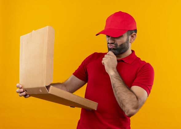 制服留着胡须的送货员穿着红色制服 戴着帽子 手里拿着打开的披萨盒 困惑地看着它 非常焦虑地站在橙色的墙上帽子男人迷茫