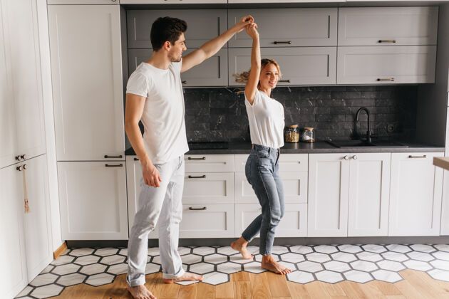 金发穿着牛仔裤的时髦女孩早上和丈夫跳舞轻松的年轻人在厨房里玩的室内写真在一起乐趣家庭