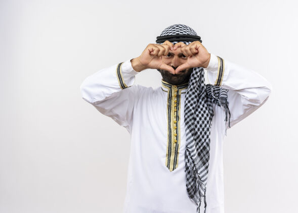 手指身着传统服装的阿拉伯男子站在白色的墙上 用手指做着心形手势 微笑着看着手指传统通过手势