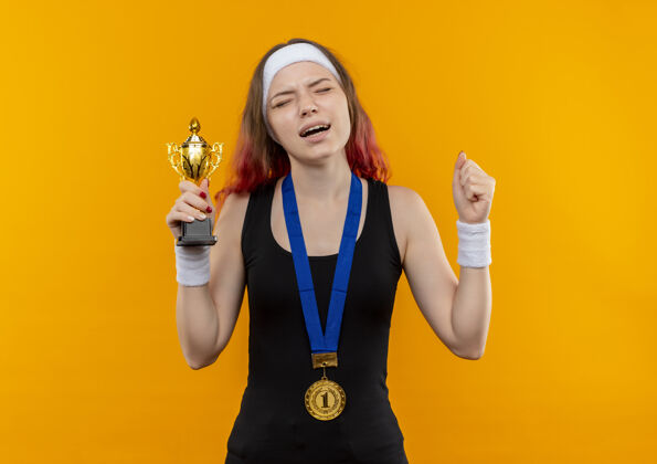 表情一位身穿运动服 脖子上挂着金牌的年轻女子举起拳头 手里拿着奖杯 脸上带着恼怒的表情站在橙色的墙上围着女性金牌