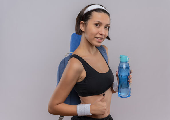 微笑年轻的健身女士穿着运动服 背着背包和瑜伽垫 手里拿着一瓶水 微笑着竖起大拇指站在白色的墙上年轻人运动站着