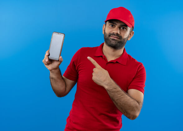 指尖一个留着胡须 身穿红色制服 戴着帽子的送货员站在蓝色的墙上 用手指着他的智能手机手指男人制服