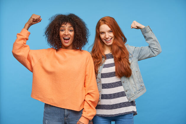 快乐快乐迷人的女士们开心地看着 开心地笑着 举起双手 同时展示强壮的肱二头肌 隔离在蓝色的墙壁上卷发毛衣女性