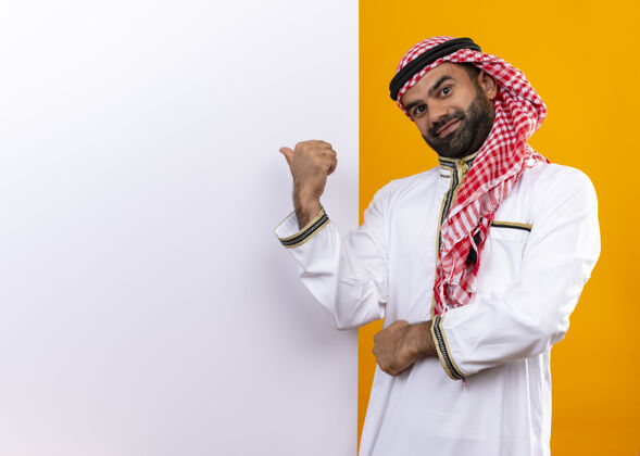 指向身着传统服装的阿拉伯商人站在空白的广告牌旁 手指指着广告牌 脸上带着自信的微笑 越过橙色的墙壁站脸阿拉伯语