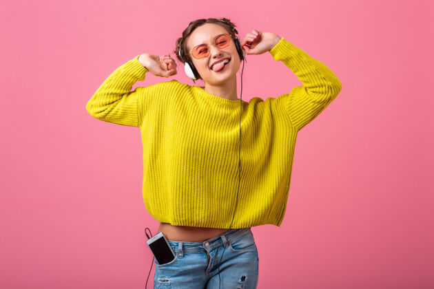 快乐迷人的快乐有趣的女人戴着耳机听音乐穿着时髦的五颜六色的服装隔离在粉红色的墙上 穿着黄色的毛衣和太阳镜 玩得很开心时尚明亮音乐