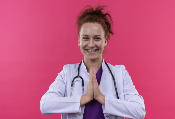 医生年轻的女医生穿着白大褂 手持听诊器 双手并拢 摆出一副非常优雅的姿势 站在粉色的墙上 微笑着表达感激和积极的情绪站着年轻外套