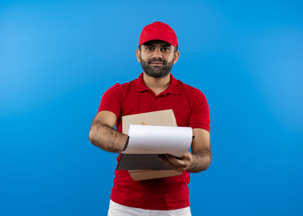 男人留着胡须的送货员身穿红色制服 戴着帽子 拿着盒子和剪贴板 上面有空白页 要求签名 微笑着站在蓝色的墙上页空白制服