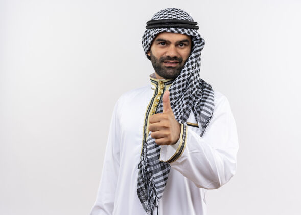 拇指身着传统服装的阿拉伯男子微笑着竖起大拇指站在白墙上穿站阿拉伯语