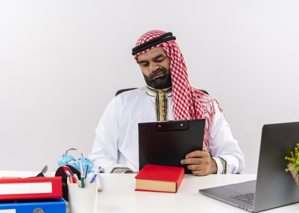文件身着传统服装的阿拉伯商人坐在桌子旁 拿着剪贴板看文件 不高兴地在办公室工作坐着商务人士阿拉伯语