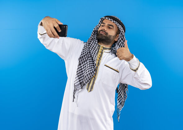 采取身着传统服装的阿拉伯男子用智能手机自拍 站在蓝色墙壁上竖起大拇指使用自拍男人
