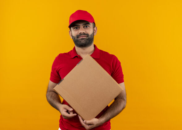 拿着有胡子的送货员穿着红色制服 戴着帽子 脸上带着微笑站在橙色的墙上拿着盒子包装胡子看