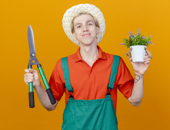 年轻高兴的年轻园丁男子穿着连体衣和帽子举行树篱修剪花盆请快乐