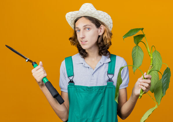 篱笆年轻的园丁穿着连体衣戴着帽子拿着植物和树篱剪穿年轻人站着