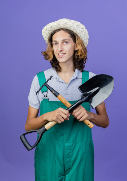 年轻人年轻的园丁 穿着连体衣 戴着帽子 手里拿着小耙子和铲子男人穿迷你