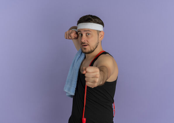 运动员戴着头带 肩上拿着毛巾的年轻健身男子站在紫色的墙上 向前伸出拳头教练拳头人