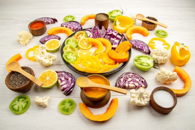 盘子俯瞰切碎的蔬菜和水果南瓜柿子红色卷心菜柠檬绿色西红柿花椰菜黄色甜椒放在圆盘子里香料放在小碗里放在白色的桌子上卷心菜胡椒小的