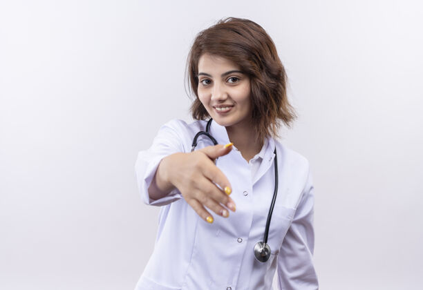 年轻身穿白大褂的年轻女医生带着听诊器微笑着自信地招呼站在白墙上的一只手听诊器问候手