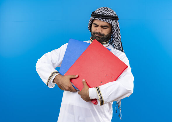 阿拉伯语身着传统服装的阿拉伯商人站在蓝色的墙上 手里拿着两个文件夹 看上去很困惑商务人士文件夹传统