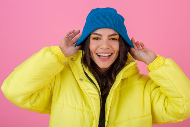 针织一幅迷人微笑的时尚女人的画像 穿着黄色的彩色冬季羽绒服 戴着蓝色的针织帽子 穿着暖和的衣服 在粉色的墙上摆姿势 时尚潮流时尚乐趣天气
