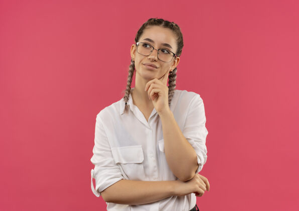 姿势戴着眼镜 梳着辫子 穿着白衬衫的年轻女学生站在粉红色的墙上 手放在下巴上 困惑地看着一边衬衫人手势