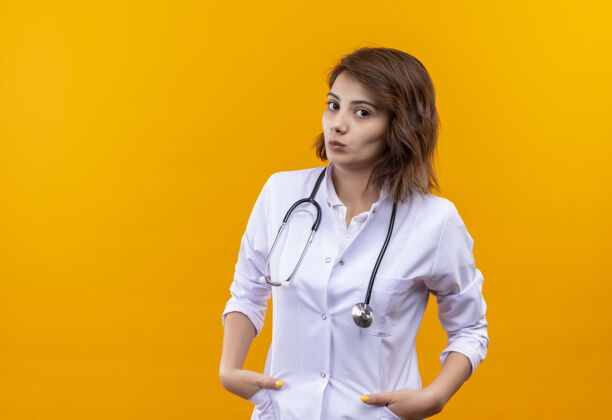 女人身穿白大褂 手持听诊器的年轻女医生站在那里 双手插在口袋里 严肃的脸越过橘黄色的墙壁外套听诊器严肃