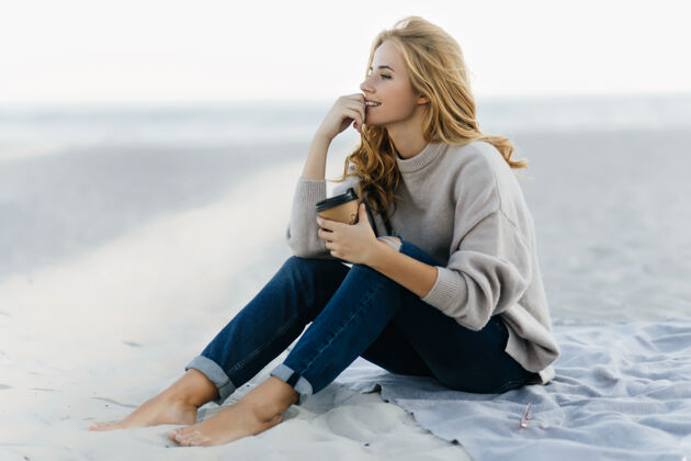周末沉思的盲人妇女穿着牛仔裤坐在沙滩上看海放松的白人妇女在海滩喝咖啡的户外肖像户外放松风