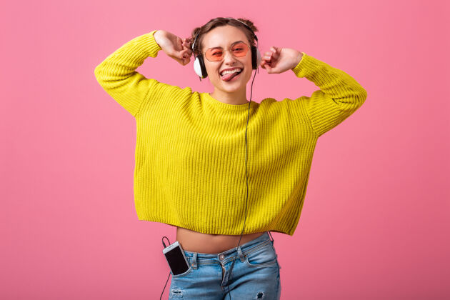 学生迷人的快乐有趣的女人戴着耳机听音乐穿着时髦的五颜六色的服装隔离在粉红色的墙上 穿着黄色的毛衣和太阳镜 玩得很开心聆听运动有趣