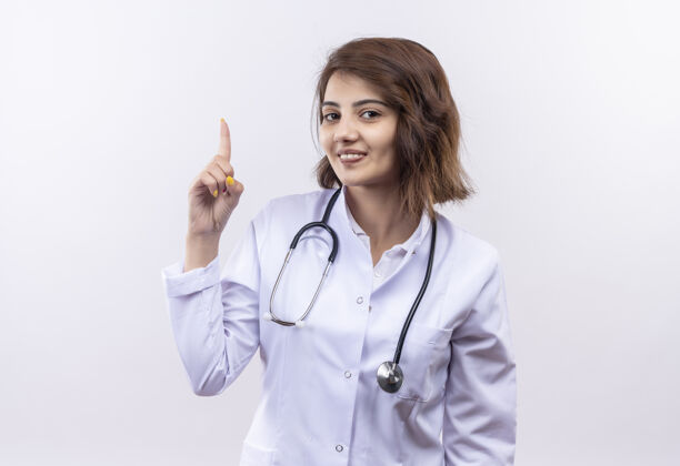 微笑身穿白大褂的年轻女医生带着听诊器微笑着自信地站在白墙上展示着有着新的伟大想法的食指女人医生目录