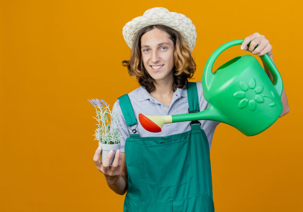 穿年轻的园丁穿着连体衣戴着帽子拿着浇水罐植物举行微笑