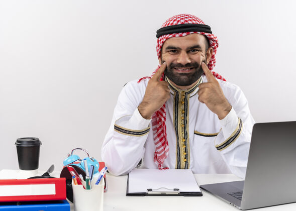 坐着身着传统服装的阿拉伯商人坐在桌旁 用手指指着笔记本电脑 微笑着假装在办公室工作指阿拉伯语假的