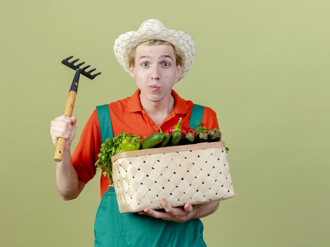 帽子年轻的园丁穿着连体衣 戴着帽子 手里拿着装满蔬菜和小耙子的板条箱 站在灯光背景下困惑地看着摄像机年轻站着全