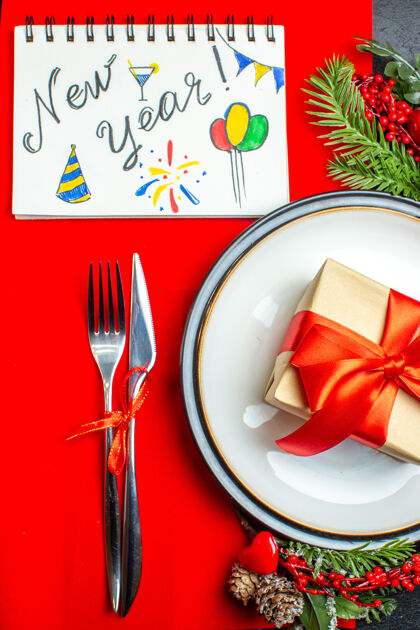 配件晚餐盘子的俯视图 上面有礼物和冷杉枝餐具套装装饰配件针叶树圆锥体下一个笔记本 红色餐巾上有新年文字和图画书写冷杉针叶树