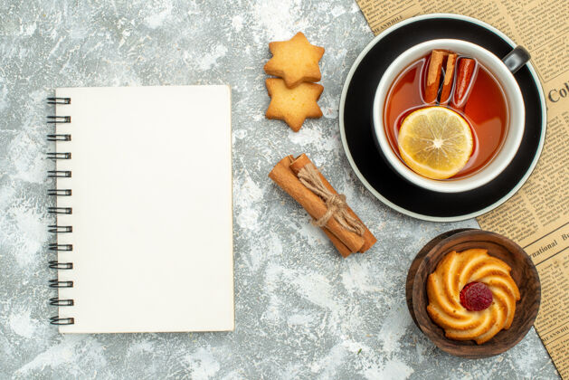 碗俯视图一杯茶 柠檬片和肉桂棒放在灰色表面的报纸笔记本饼干上笔记本香料茶