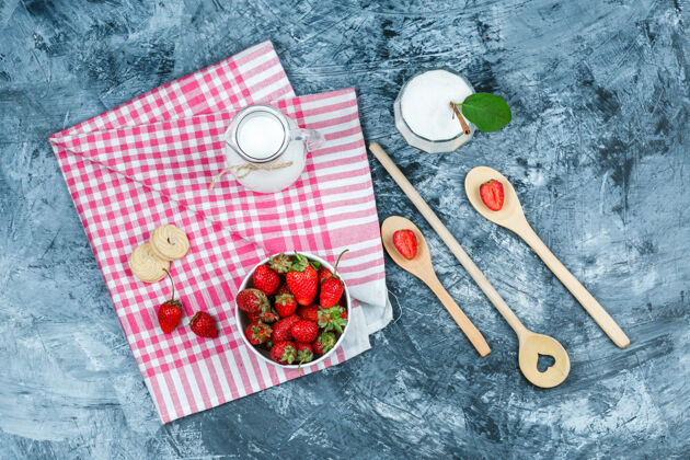 毛巾把一碗草莓和一壶牛奶平放在红方格毛巾上 用木勺子和一杯酸奶放在深蓝色大理石表面水平草莓配料水果