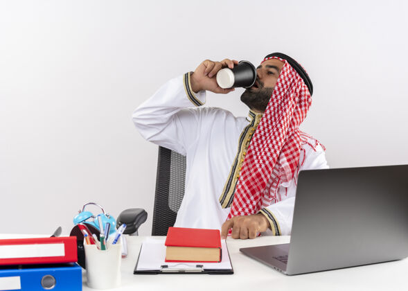 喝身着传统服装的阿拉伯商人坐在桌旁 拿着笔记本电脑喝着咖啡在办公室工作坐阿拉伯语传统