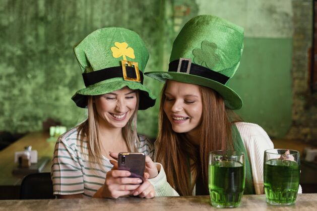 帽子快乐的女性朋友一起在酒吧用智能手机庆祝圣帕特里克节场合绿色三叶草