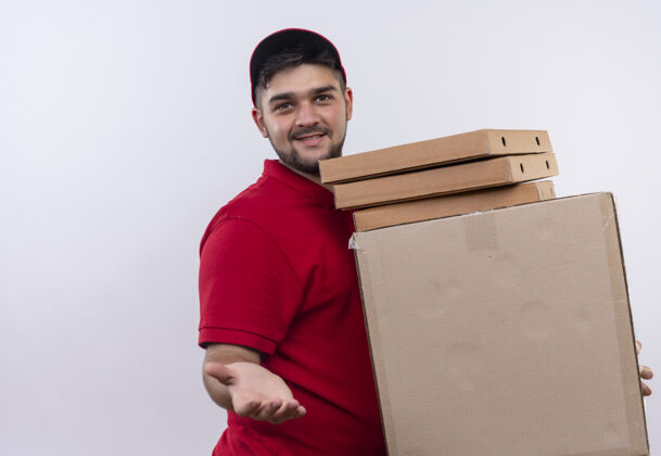 立场年轻的送货员身着红色制服 戴着帽子 手里拿着一叠盒子 友好地致意致意年轻制服微笑
