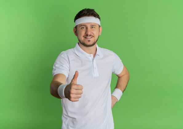 脸身穿白衬衫 头箍 面带微笑 竖起大拇指站在绿色墙壁上的年轻健身男子年轻人表演微笑