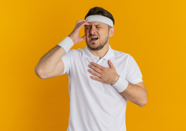 人身穿白衬衫 头箍不适的年轻健身男子站在橙色的墙上 头痛得厉害头带看运动