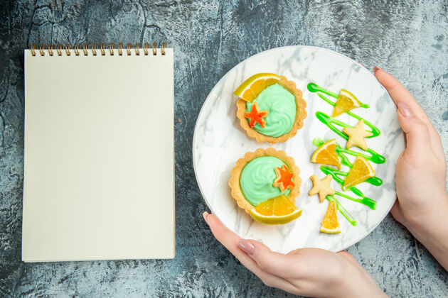 切片顶视图小馅饼与绿色糕点奶油和柠檬片在妇女手中的盘子笔记本在灰色的桌子上食物手盘子