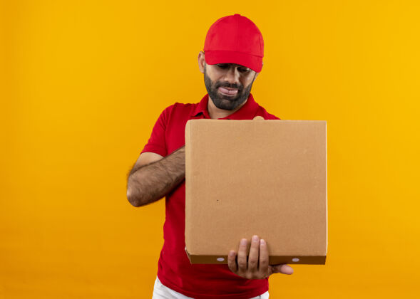 披萨一个留着胡须的送货员 穿着红色制服 戴着帽子 手里拿着打开的披萨盒 站在橘色的墙上 一脸滑稽地看着它男人站着胡子