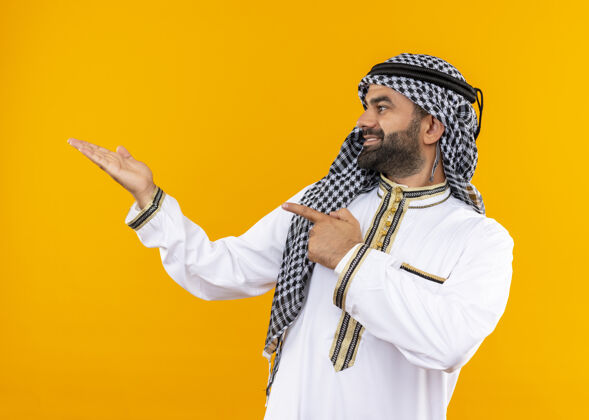 商人身着传统服装的阿拉伯商人一边看一边用手指和胳膊指着站在橙色墙上的一边传统阿拉伯语手指