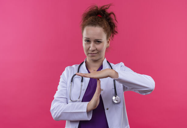 听诊器年轻的女医生穿着白色外套 手持听诊器 看起来很累 很无聊 双手站在粉红色的一边 做着暂停的手势站外套手势